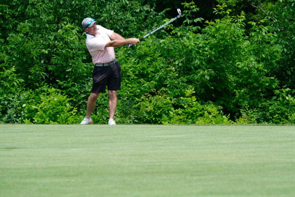A man wearing a pink golf shirt and black shorts swings his golf club. He is standing on green grass with lush green trees behind him