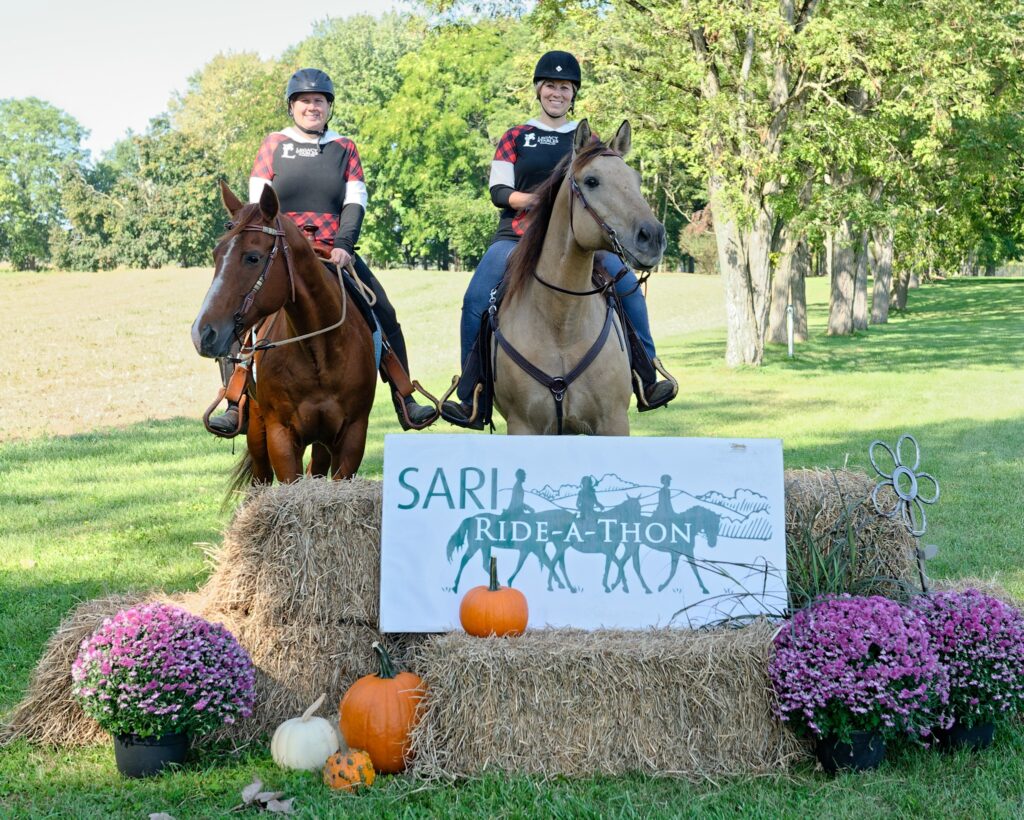 Two women mounted on horses pose behind a stack of hay bales displaying a sign that says SARI Ride a Thon and decorated with pumpkins and mums flowers.