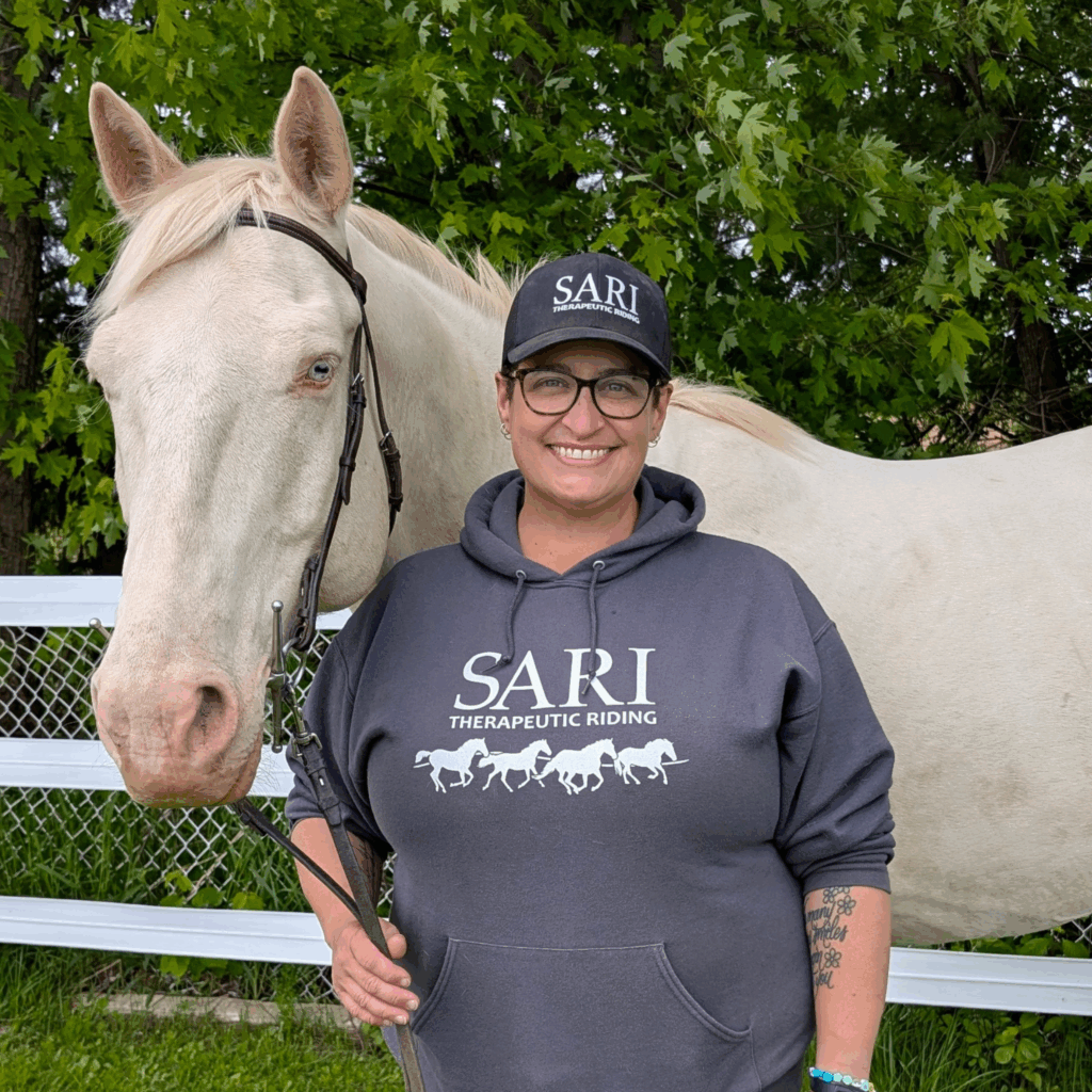A woman with dark hair, wearing a black trucker hat and a gray hoodie with the SARI logo on the front, stands next to a white horse. The woman is holding a lead rope and smiling at the camera