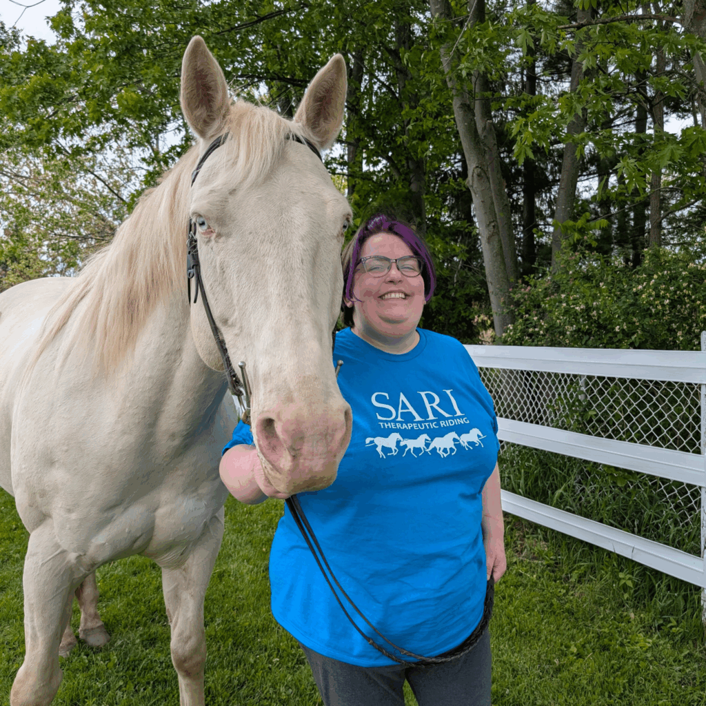 A smiling person with brown and purple hair, wearing a blue shirt with the SARI logo on the front, stands next to a white horse.