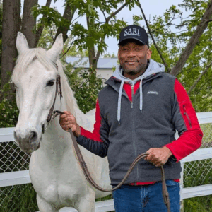 A man wearing a grey hoodie under a black and red jacket and a black trucker hat with the SARI logo on it stands next to a white horse.