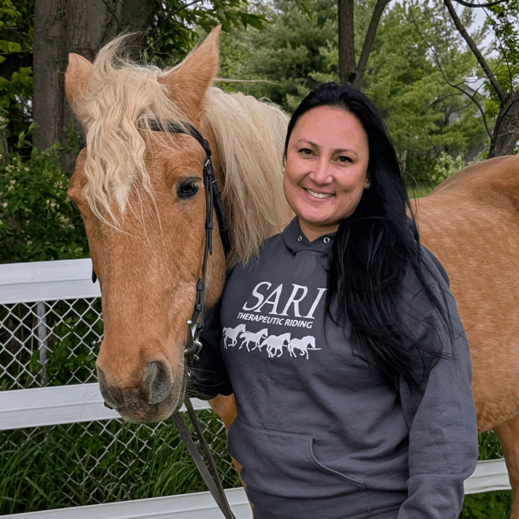 A smiling woman with long dark hair and wearing a grey hoodie with the SARI logo on it stands next to a palomino horse.