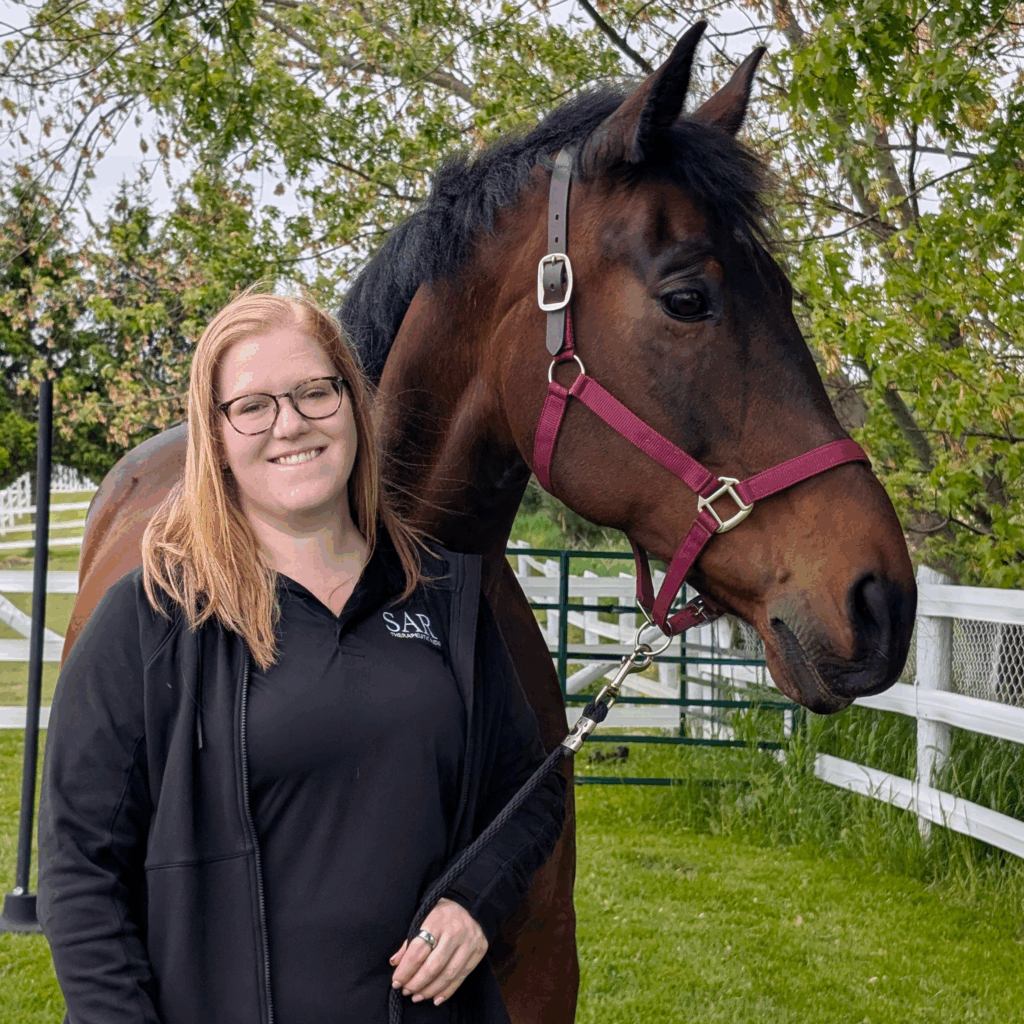 A smiling woman with long strawberry blonde hair stands next to a dark brown horse.