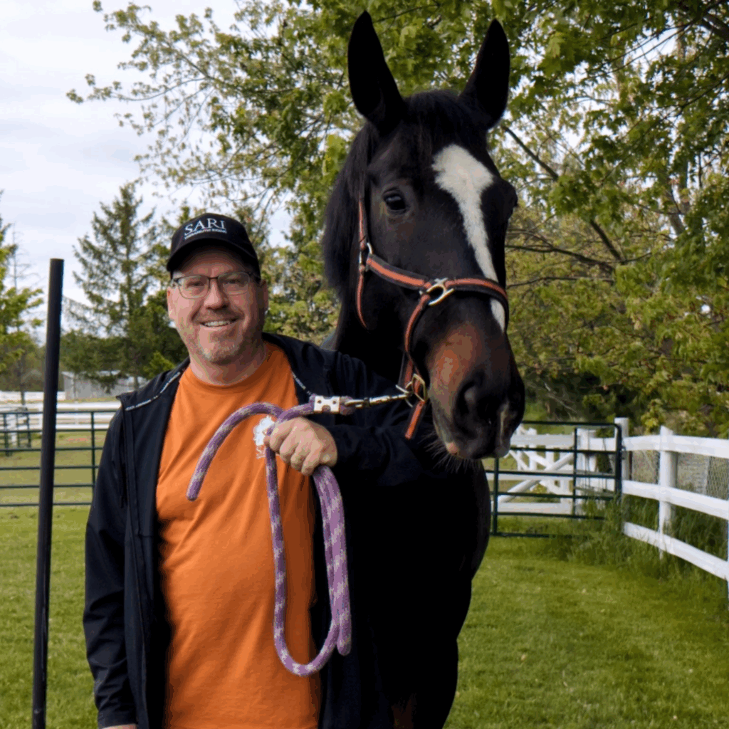 A smiling man wearing a bright orange t-shirt stands next to a tall, dark brown horse