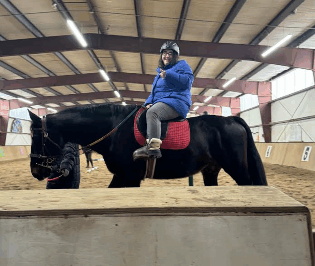 A young woman in a blue puffer jacket, blue jeans, glasses, and a black riding helmet sits in a saddle on top of a dark brown horse. The woman is smiling at the camera and giving a thumbs up. The horse is facing to the left with its head low, and looks calm.