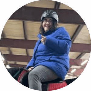 A young woman in a blue puffer jacket, blue jeans, glasses, and a black riding helmet sits in a saddle. She is smiling at the camera and giving a thumbs up.