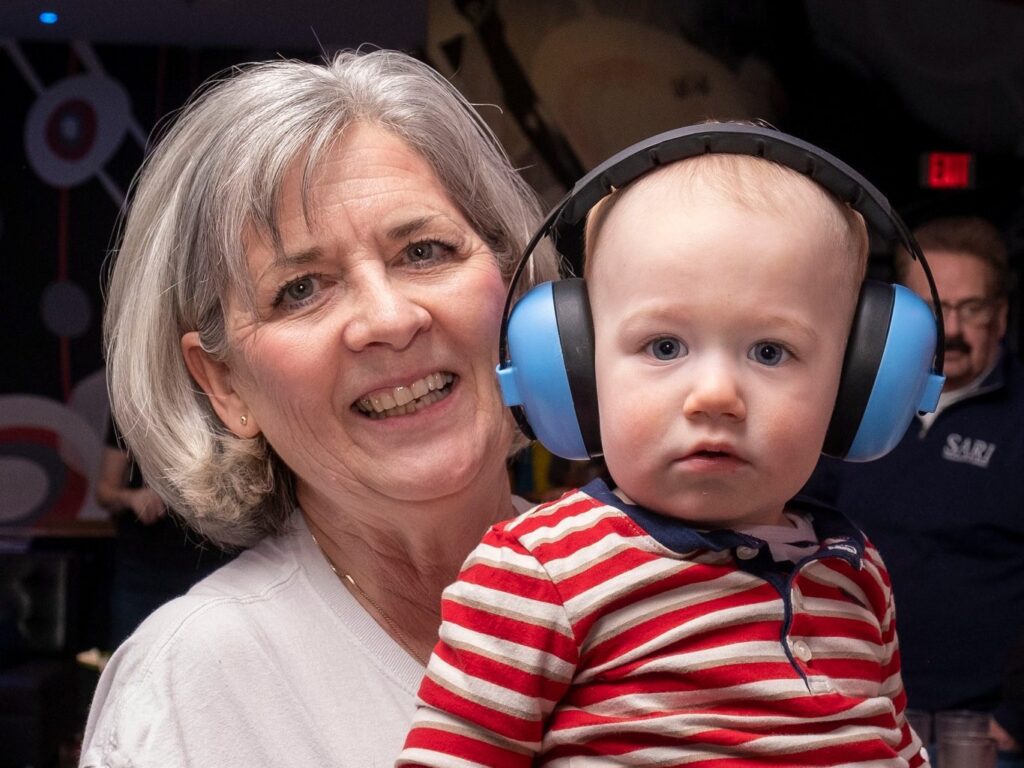 A woman with short grey hair holds a young child who is wearing large blue headphones on his head