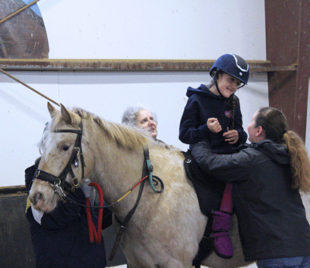 A girl sitting sideways on a horse. On either side of the horse there is an adult smiling and watching the rider