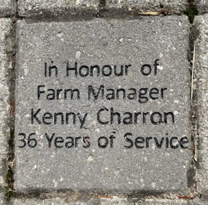 A grey brick laid into the ground that reads "In honour of Farm Manager Kenny Charon 36 years of service"