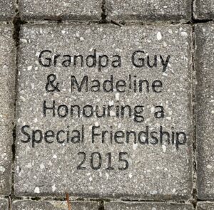 A grey brick laid into the ground that reads "Grandpa Guy and Madeline honouring a special friendship 2015"