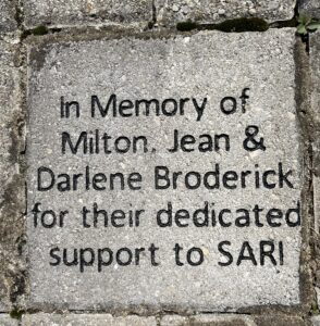 A grey brick laid into the ground that reads "In memory of Milton, Jean, and Darlene Broderick for their dedicated support to SARI"